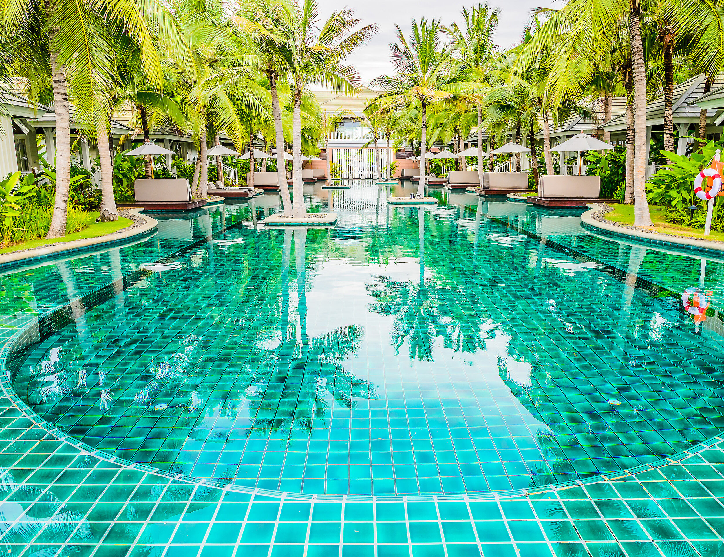 Elegant outdoor swimming pool with crystal-clear blue tiles, surrounded by palm trees, private cabanas, and lush greenery — perfect for a wedding pool party.
